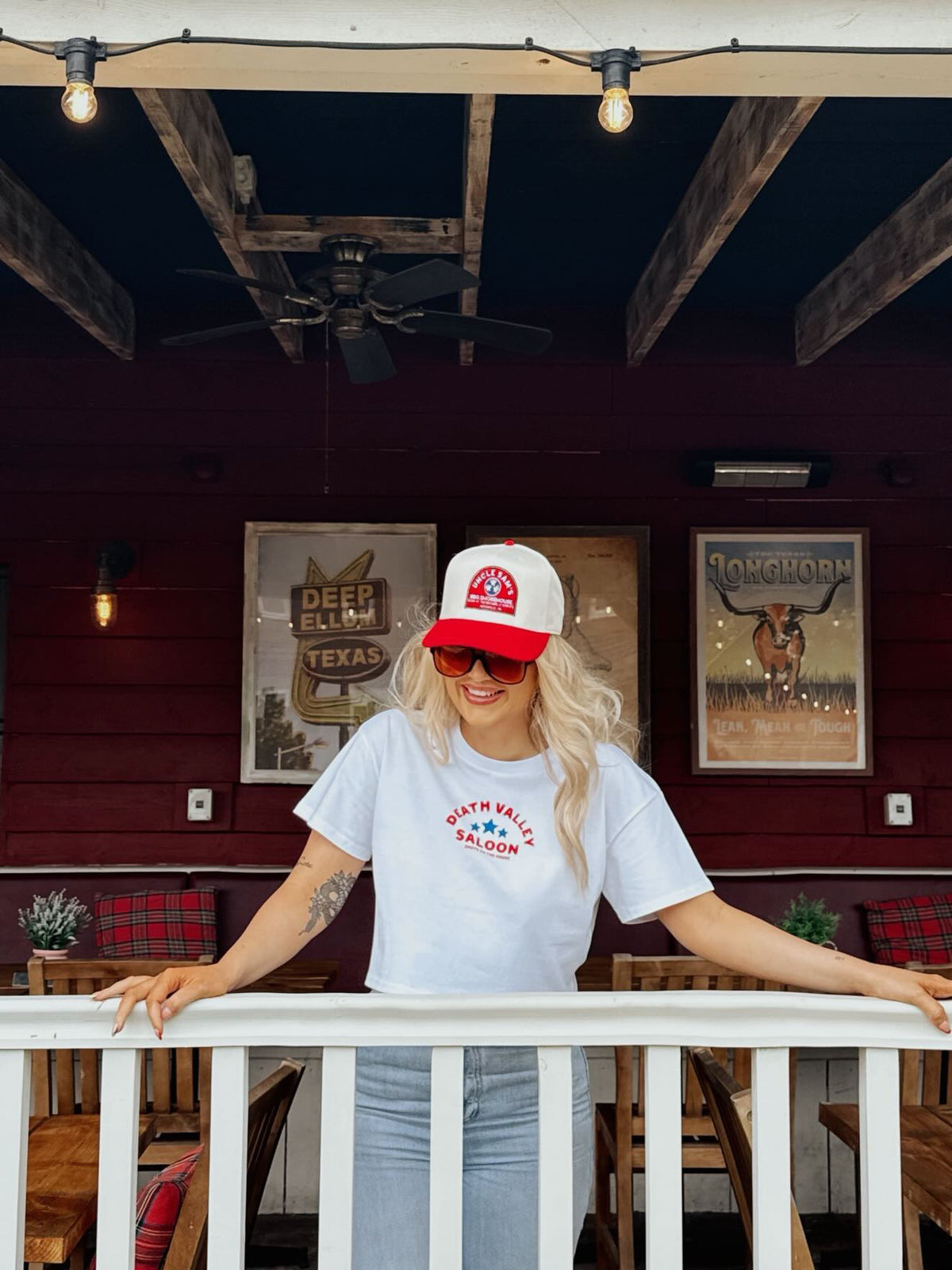 A woman stands on a porch wearing a white t-shirt with red and blue 'Sourpuss Ranch' text, a matching red and white trucker hat, and sunglasses. The background features a dark red wooden wall with rustic framed signs and hanging light bulbs.