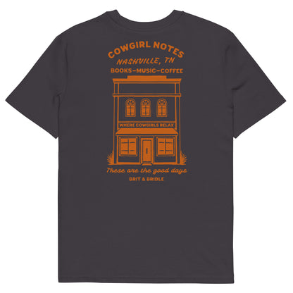 A dark charcoal gray t-shirt shown from the back, featuring a prominent orange graphic print of a vintage building with the text 'COWGIRL NOTES NASHVILLE, TN BOOKS-MUSIC-COFFEE' and 'These are the good days BIT & BRUISE'.