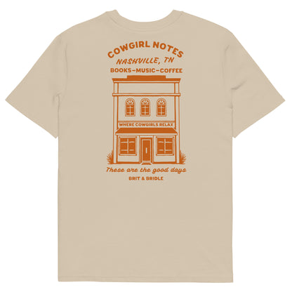 A beige short-sleeve t-shirt shown from the back, featuring an orange graphic print of a vintage storefront building with the text 'COWGIRL NOTES NASHVILLE, TN BOOKS-MUSIC-COFFEE' and 'These are the good days BRIT & BRIDLE' printed below.