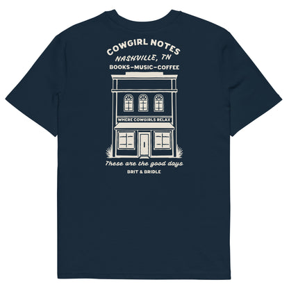 A navy blue short-sleeve t-shirt with a white graphic print on the front depicting a storefront and text reading 'COWGIRL NOTES NASHVILLE, TN BOOKS - MUSIC - COFFEE' and 'These are the good days BRIT & BRIDLE'.