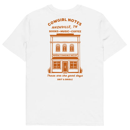 A white short-sleeve t-shirt with a retro-style orange graphic on the front. The graphic features a two-story building illustration with the text 'COWGIRL NOTES NASHVILLE, TN BOOKS-MUSIC-COFFEE' above it, and 'These are the good days BINT & BRIDLES' below it.